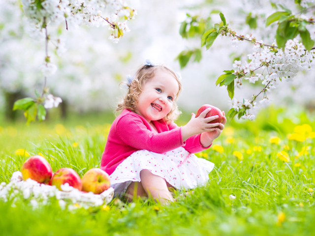Маленькая улыбающаяся девочка сидит на зеленой траве с яблоками