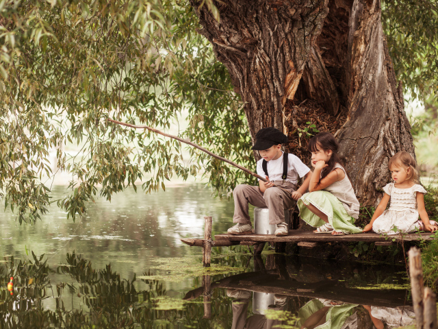 Две маленькие девочки и мальчик сидят на берегу пруда с удочкой