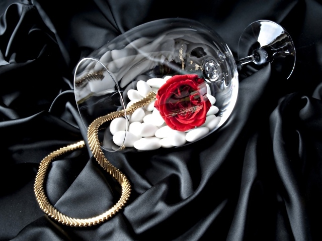 Бокал с золотой цепочкой, красной розой и белыми камнями на черном покрывале