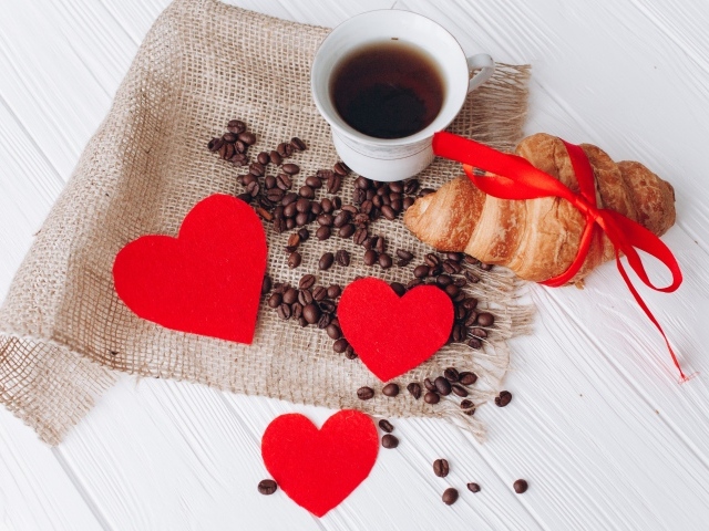 Круассан с чашкой кофе и кофейными зернами на столе с красными сердечками