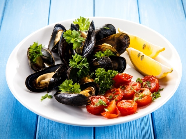 Мидии с томатами, кусочками лимона и зеленью петрушки на белой тарелке