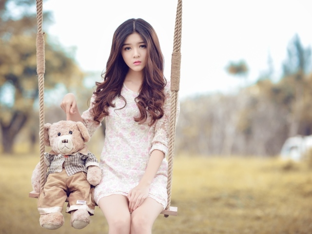 Девочка азиатка сидит на качели с плюшевым мишкой