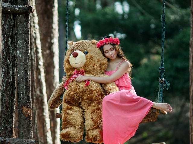 Красивая девушка азиатка в розовом платье сидит на качели с большим игрушечным медведем
