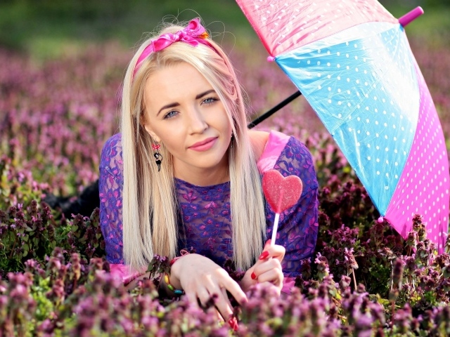 Молодая девушка блондинка лежит на поляне с сиреневыми цветами под разноцветным зонтом