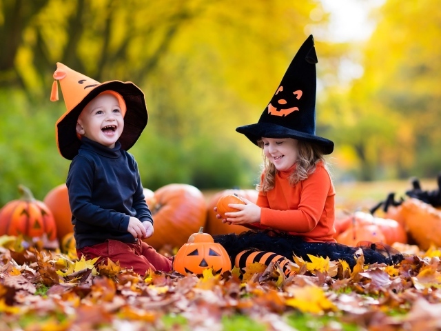 Маленькие мальчик и девочка сидят на опавших листьях с тыквами на Хэллоуин