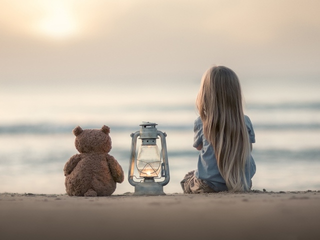 Маленькая девочка сидит на песке с плюшевым мишкой 