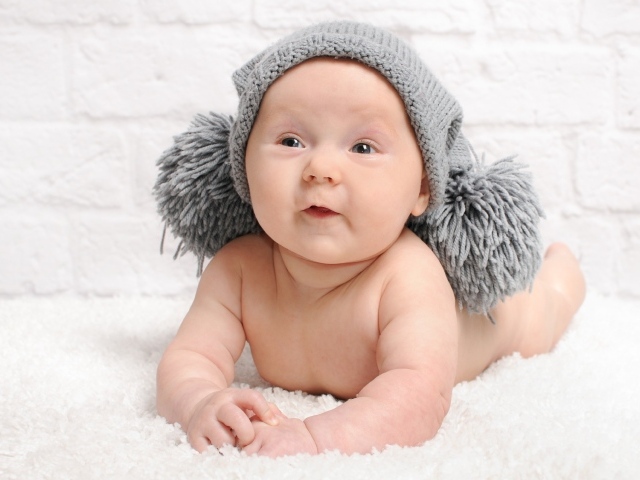 Улыбающийся грудной ребенок в серой шапке с бубонами