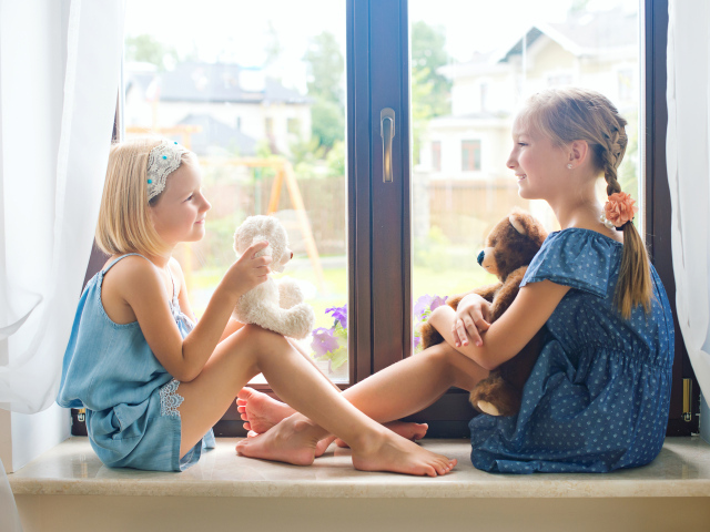 Две маленькие девочки с игрушками сидят на подоконнике у окна