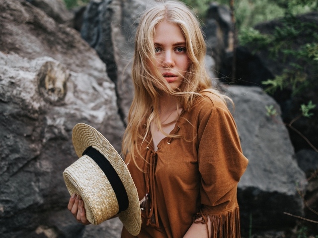 Блондинка со шляпой в руках стоит у камней