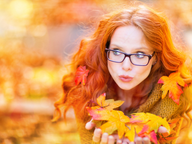 Рыжеволосая девушка в очках держит в руках опавшие листья 