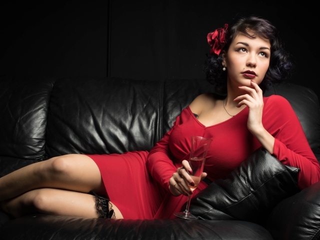 Девушка в красном платье с бокалом в руке лежит на кожаном диване