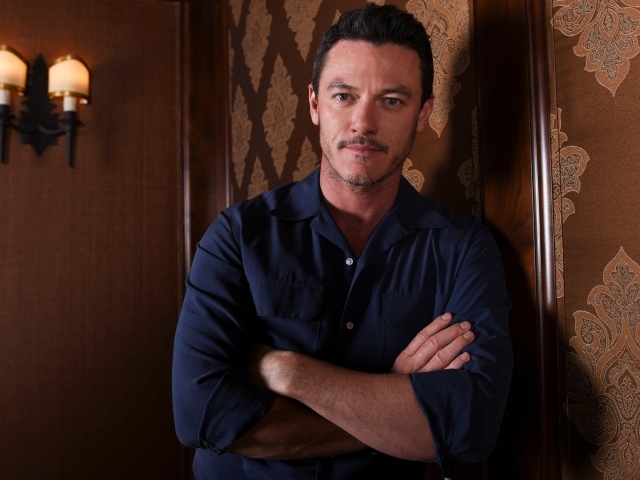 Актер Люк Эванс в рубашке стоит у стены