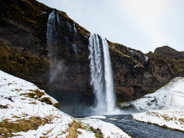Водопад стекает со скалы в реку с покрытыми снегом берегами