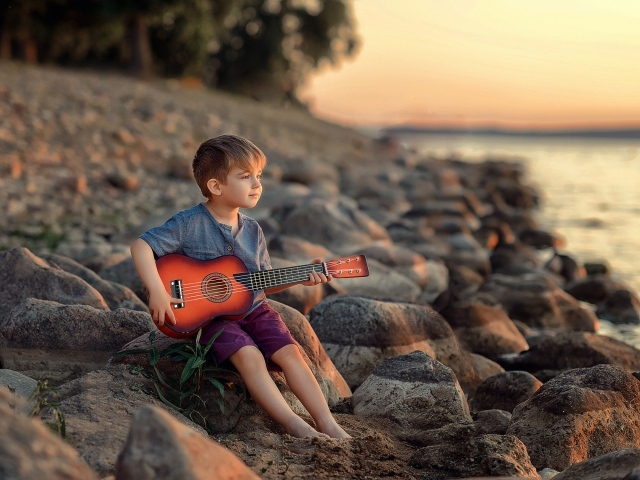 Маленький мальчик сидит с гитарой на камнях у моря 