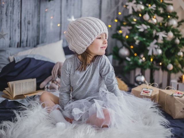 Маленькая девочка сидит на кровати у новогодней елки