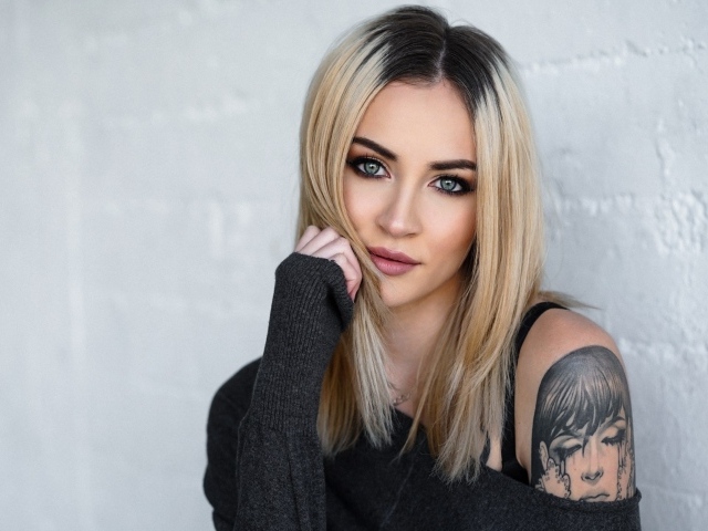 Стильная блондинка с татуировкой на плече на фоне стены