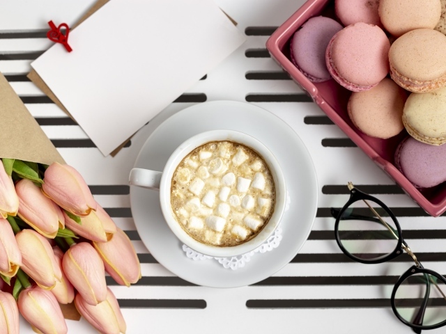 Чашка какао с маршмеллоу на столе с букетом тюльпанов, десертом макарун и очками