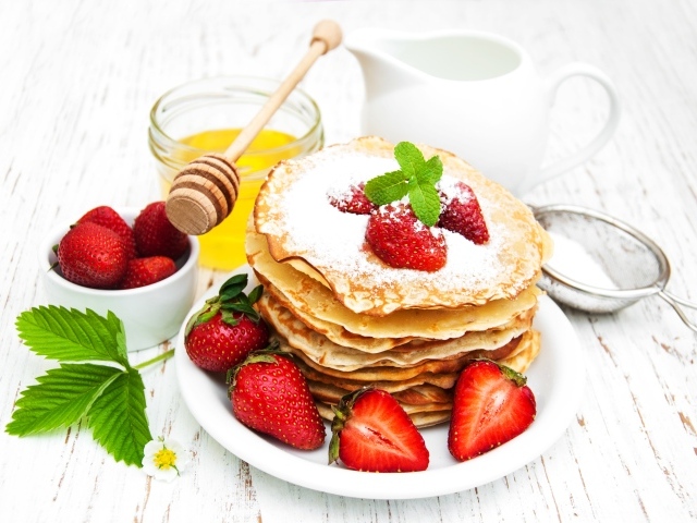 Тонкие блины с сахарной пудрой и ягодами клубники на столе с молоком и медом