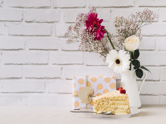 Кусок торта, подарок и букет на столе на фоне стены 
