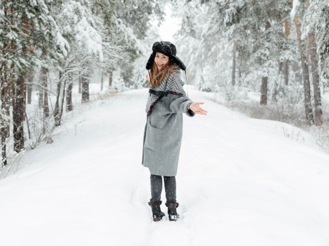 Девушка в пальто и теплой шапке гуляет в заснеженном лесу 