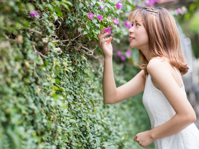 Девушка азиатка в белом платье стоит у зеленой изгороди 