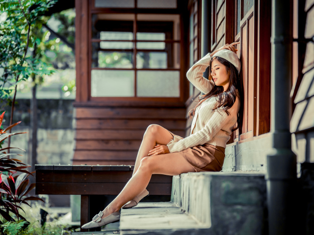 Девушка азиатка с закрытыми глазами сидит на ступеньках дома 