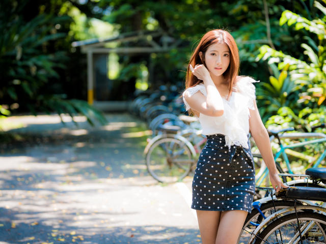 Красивая девушка азиатка в юбке в горох стоит у велосипеда 