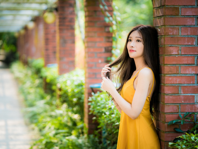 Красивая азиатка в желтом платье стоит у кирпичной колонны 