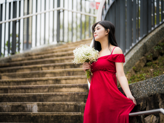 Красивая азиатка в красном платье стоит на ступеньках с букетом ландышей