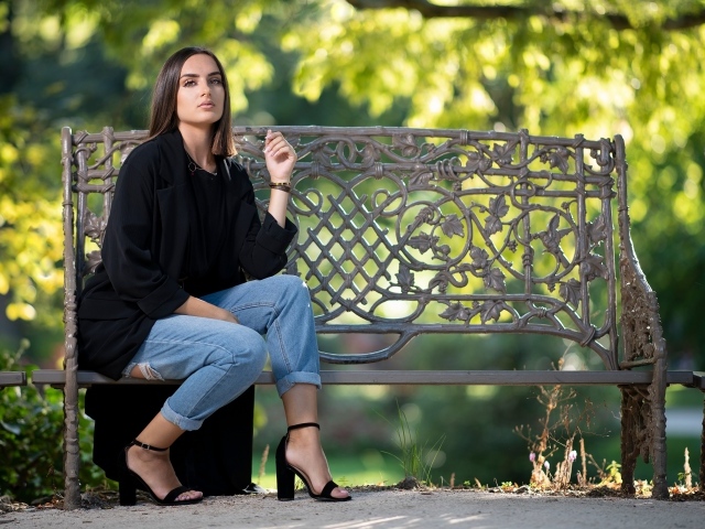 Красивая девушка в черном пальто сидит на лавочке в парке