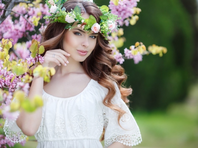 Красивая девушка в белом платье с венком на голове стоит у дерева 