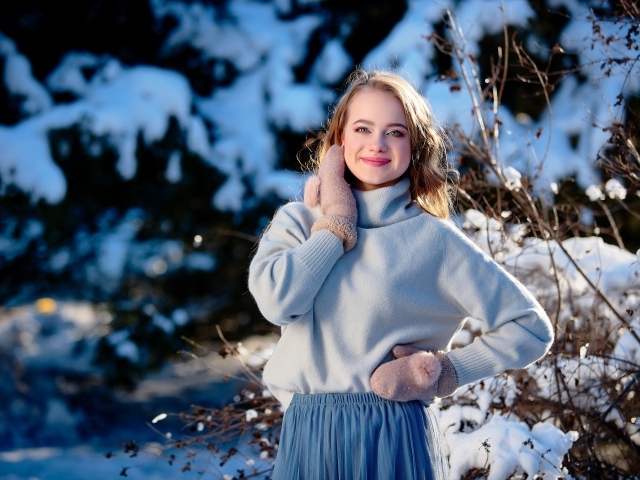 Красивая улыбающаяся девушка в варежках на фоне заснеженных деревьев 