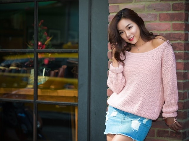 Милая азиатка в розовом свитере стоит у стены 