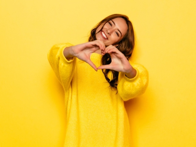 Девушка в желтом свитере показывает сердце руками