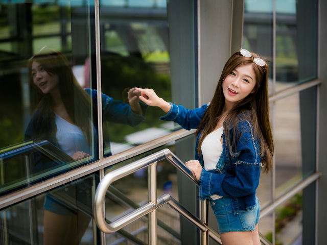 Улыбающаяся девушка в джинсовой куртке стоит у стены 