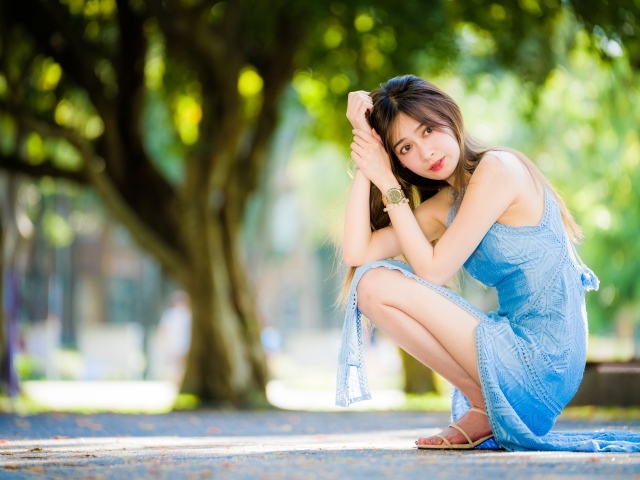 Молодая девушка азиатка в голубом платье сидит на корточках в парке 