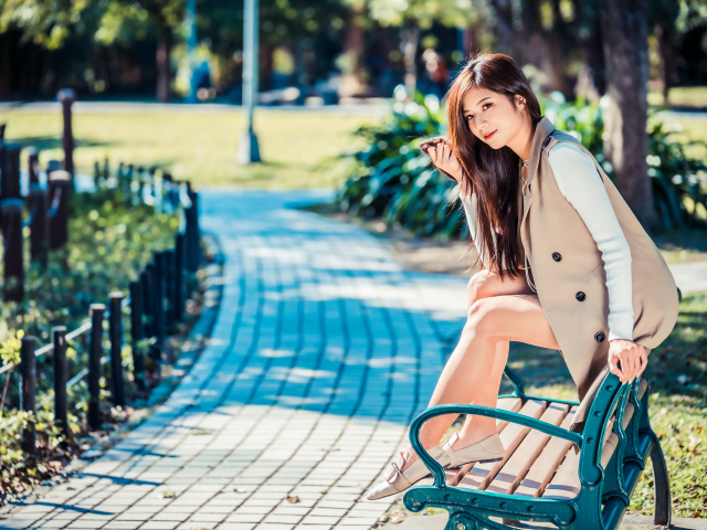 Молодая девушка азиатка сидит в пальто на лавке в парке 