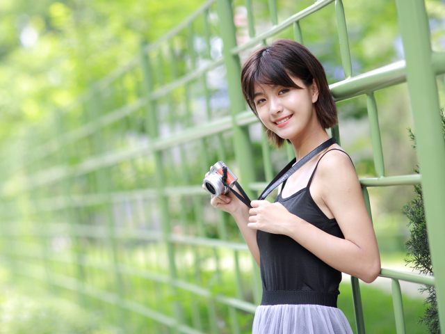 Молодая улыбающаяся азиатка с фотоаппаратом у забора 