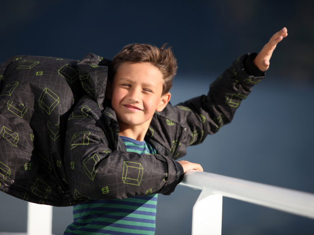 Веселый мальчик в куртке стоит на ветру в лучах солнца