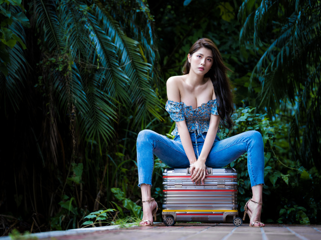 Девушка азиатка в джинсах сидит на чемодане