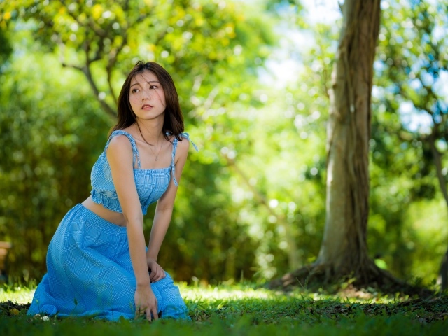 Красивая азиатка в голубом платье сидит на траве в парке 