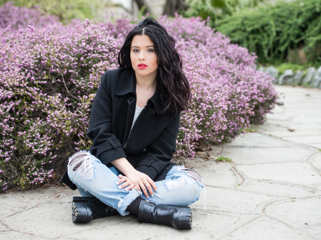 Красивая девушка в черном пальто сидит на земле у кустов