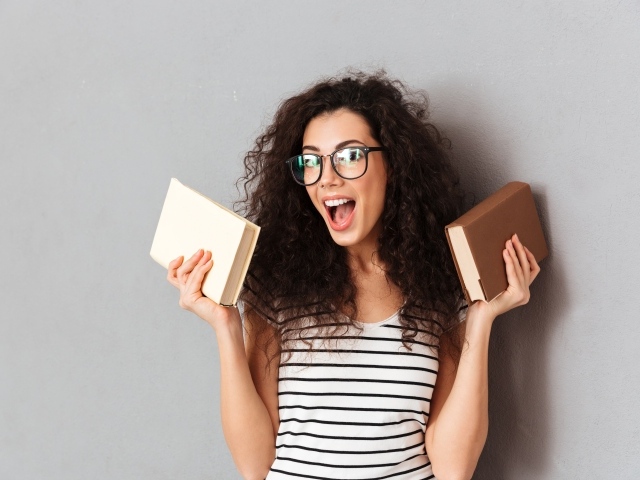 Веселая девушка в очках с книгами на фоне серой стены 