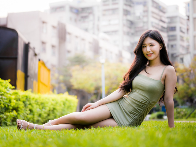 Улыбающаяся азиатка в платье сидит на зеленой траве 