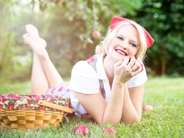 Улыбающаяся блондинка лежит на зеленой траве с корзиной яблок 