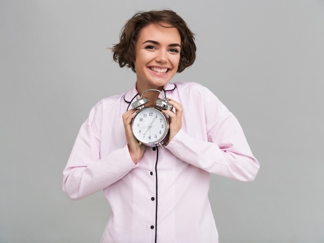 Улыбающаяся девушка в розовой рубашке с будильником в руках 