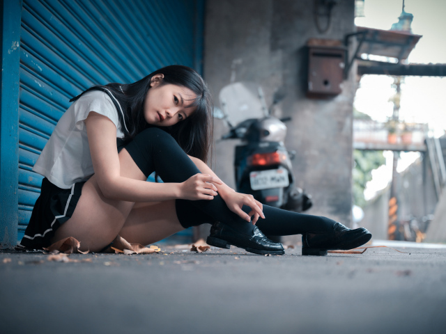 Молодая азиатка в черных чулках сидит у стены 