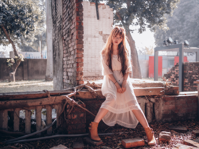 Молодая девушка азиатка в белом платье сидит на разваленной стене
