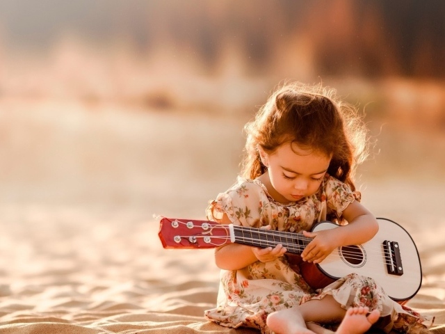 Маленькая девочка с гитарой сидит на песке 