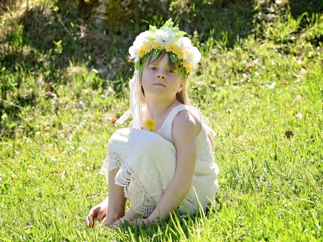 Маленькая девочка с венком на голове сидит на зеленой траве